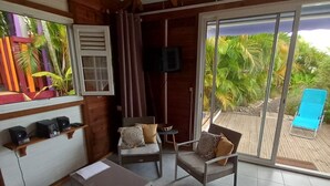Amplia habitación con ventilador de techo, televisor montado en la pared, puerta corrediza de vidrio que da a un espacio exterior con una silla lounge azul y una pequeña mesa con cojín.
