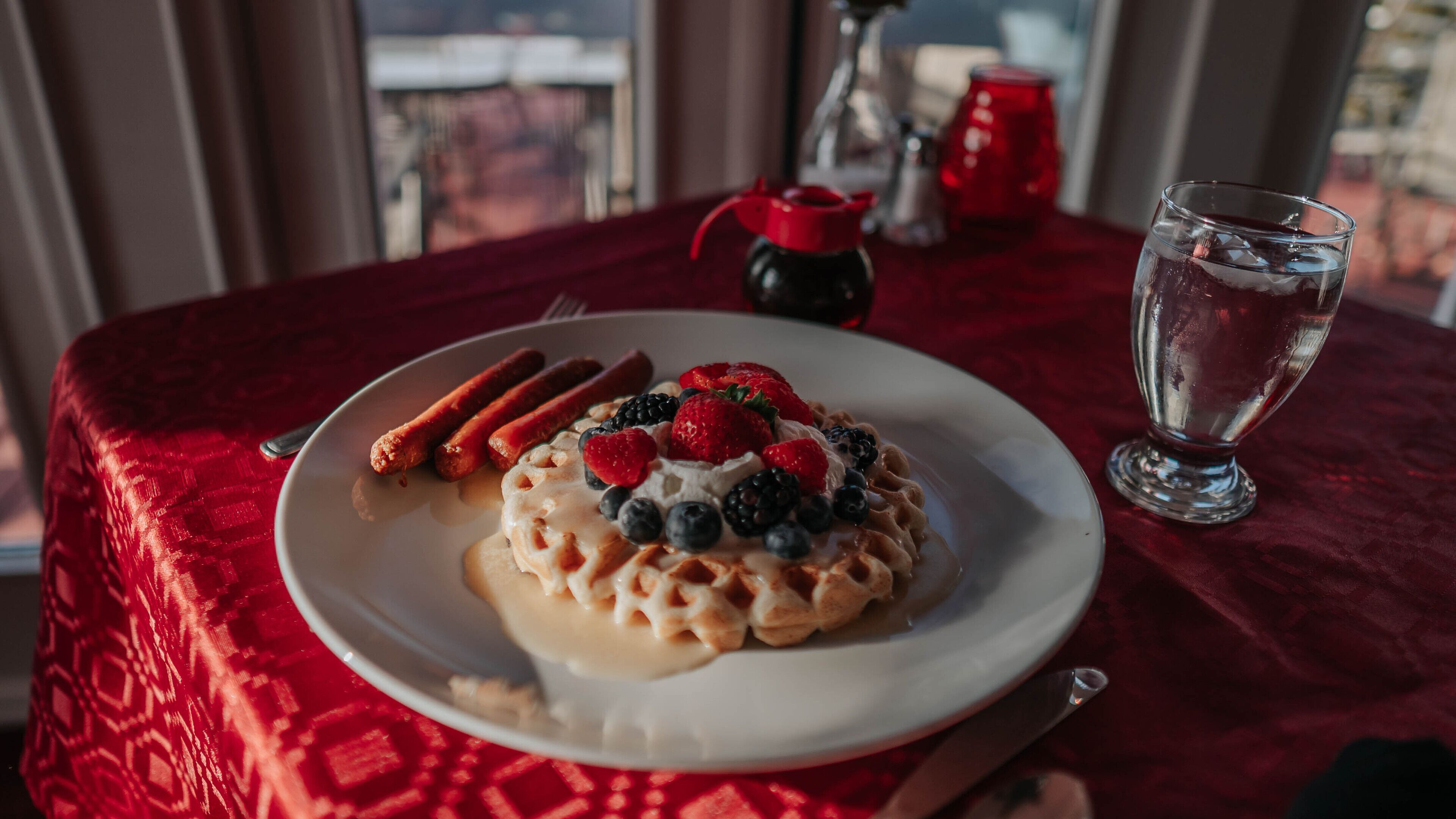 A plate of waffles topped with whipped cream, berries, and sausages on a red tablecloth.