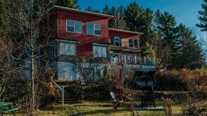 A two-story red house with a balcony, surrounded by trees and a grassy yard. - Auberge Lac du Pin Rouge (Saint-Hippolyte)