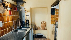 Cozinha estreita com painel de azulejos, pia e fogão. Há um tapete branco no chão e uma cadeira de madeira com um saco de batatas fritas.