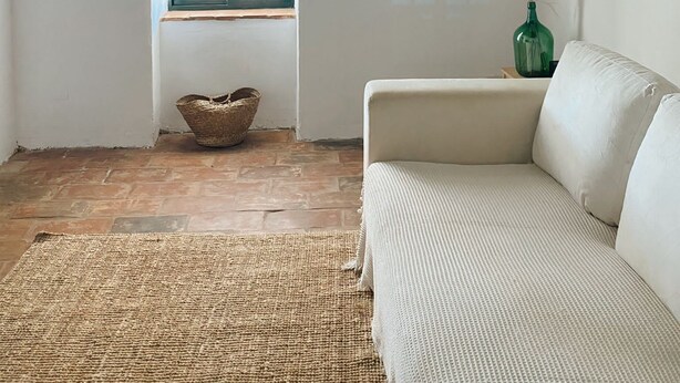 Um quarto com um sofá branco, um tapete tecido, uma janela com persianas azuis, uma cesta de vime e um vaso com plumas de pena.