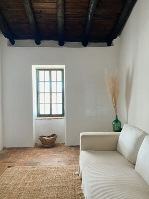 Een kamer met een witte bank, een geweven vloerkleed, een raam met blauwe luiken, een rieten mand en een vaas met pluisgras.