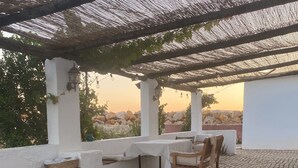 Um pátio externo com mesa, cadeiras, uma pérgula e vista para a paisagem.