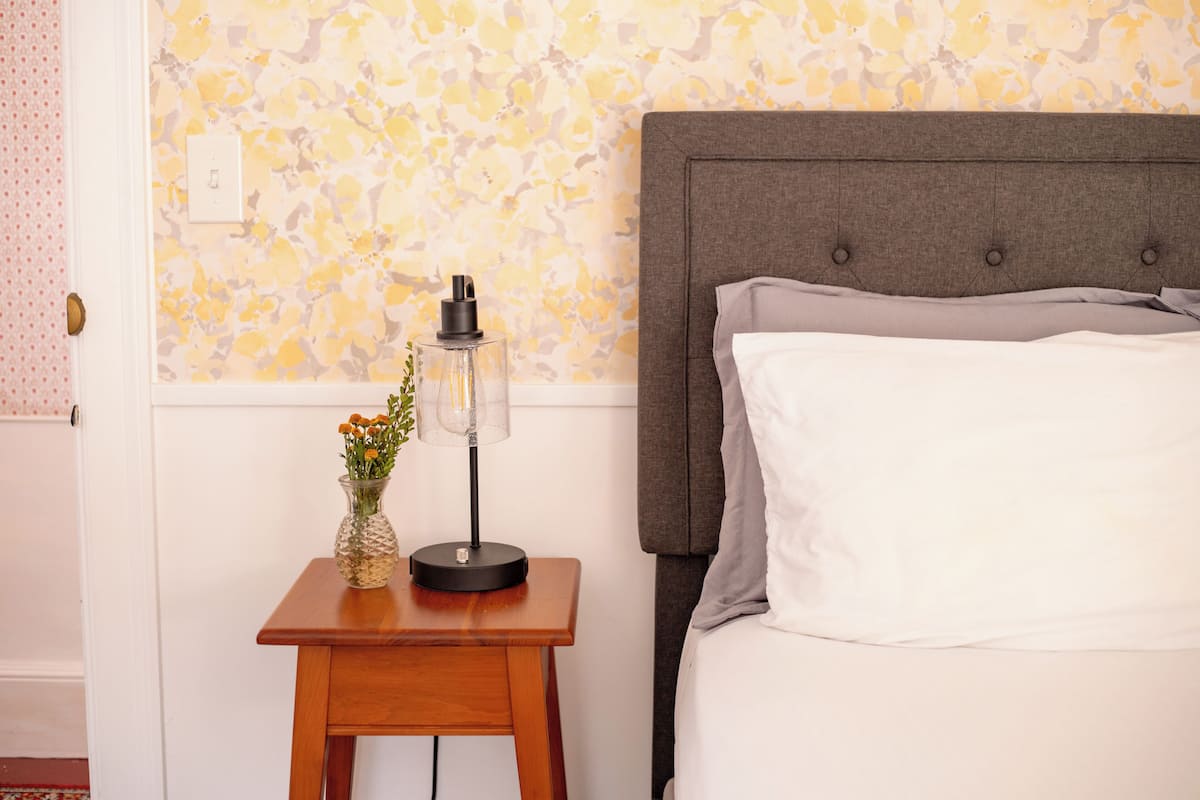 Una cama bien hecha con cabecera gris, una mesita de noche de madera con lámpara y un florero, y un papel tapiz estampado al fondo.