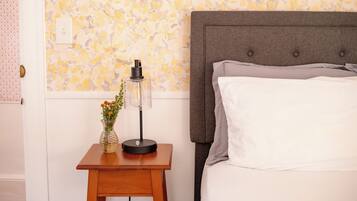 Una cama bien hecha con cabecera gris, una mesita de noche de madera con lámpara y un florero, y un papel tapiz estampado al fondo.