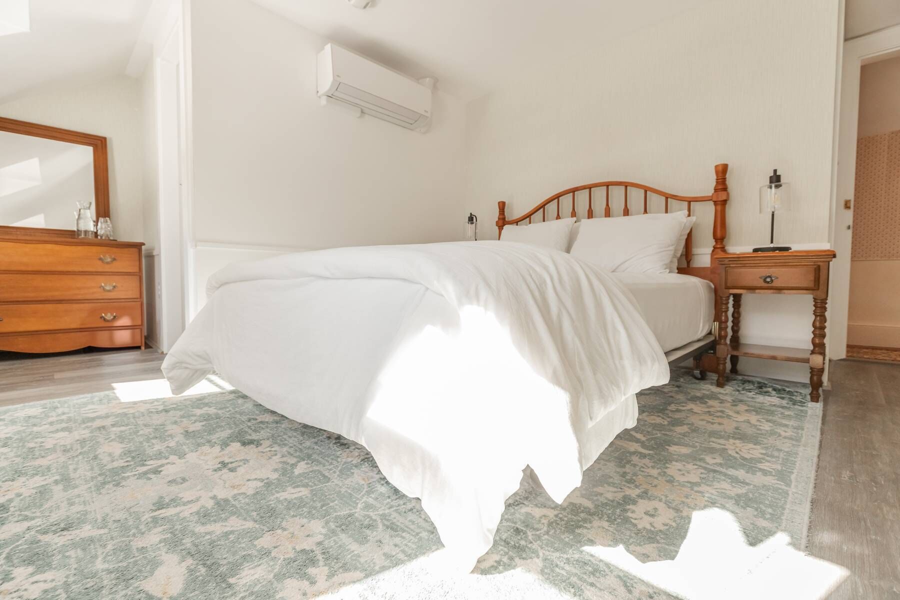 Una cama bien hecha con cabecera de madera, una mesita de noche con lámpara y un aparador con cajones.