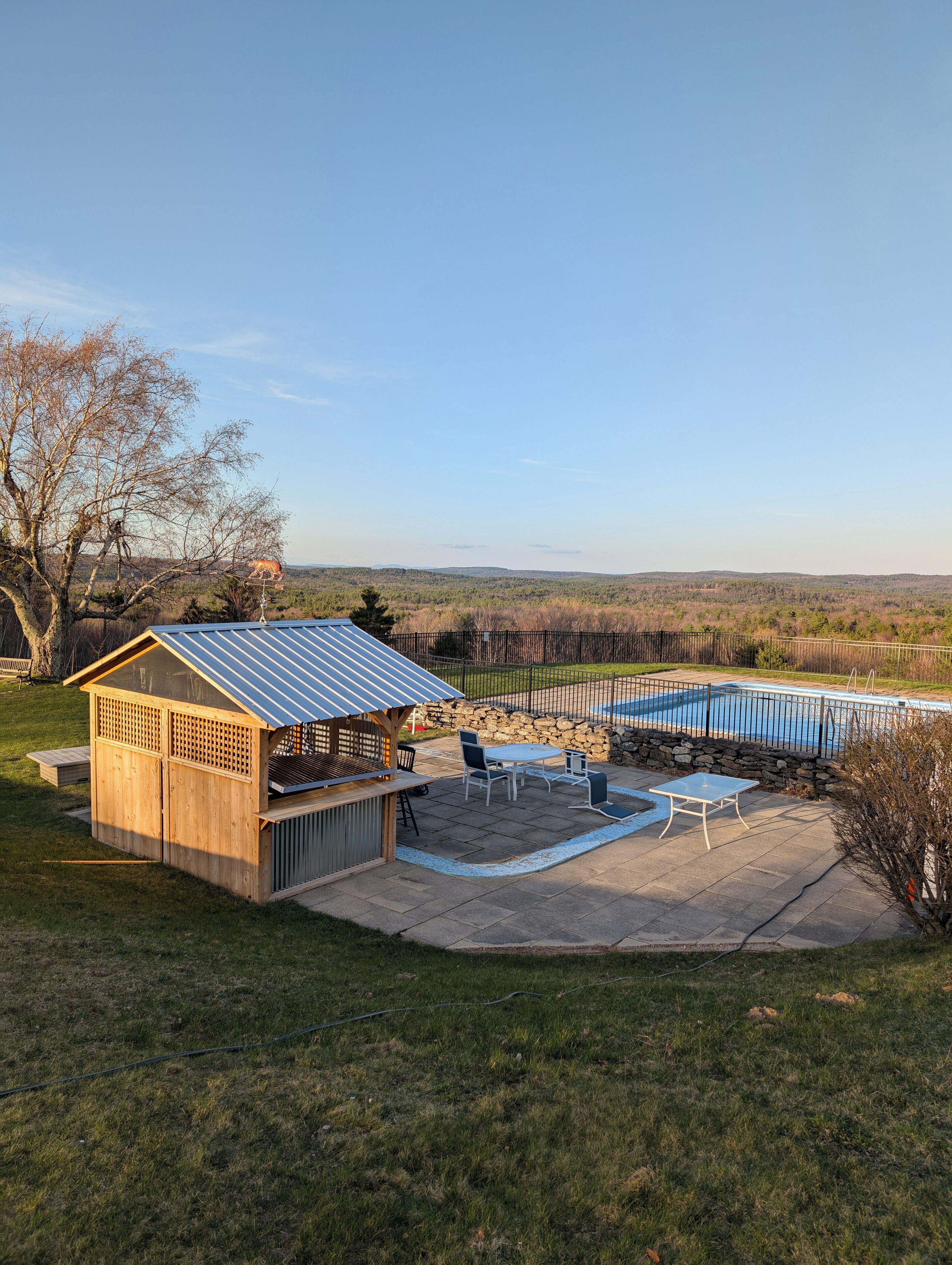 A backyard with a wooden barbecue area, a stone wall, a swimming pool, and a view of the surrounding landscape.