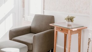 A neutral-toned armchair and a small wooden table with a glass top, positioned in a bright room with white walls and a window. - Blue Bear Inn (Francestown)