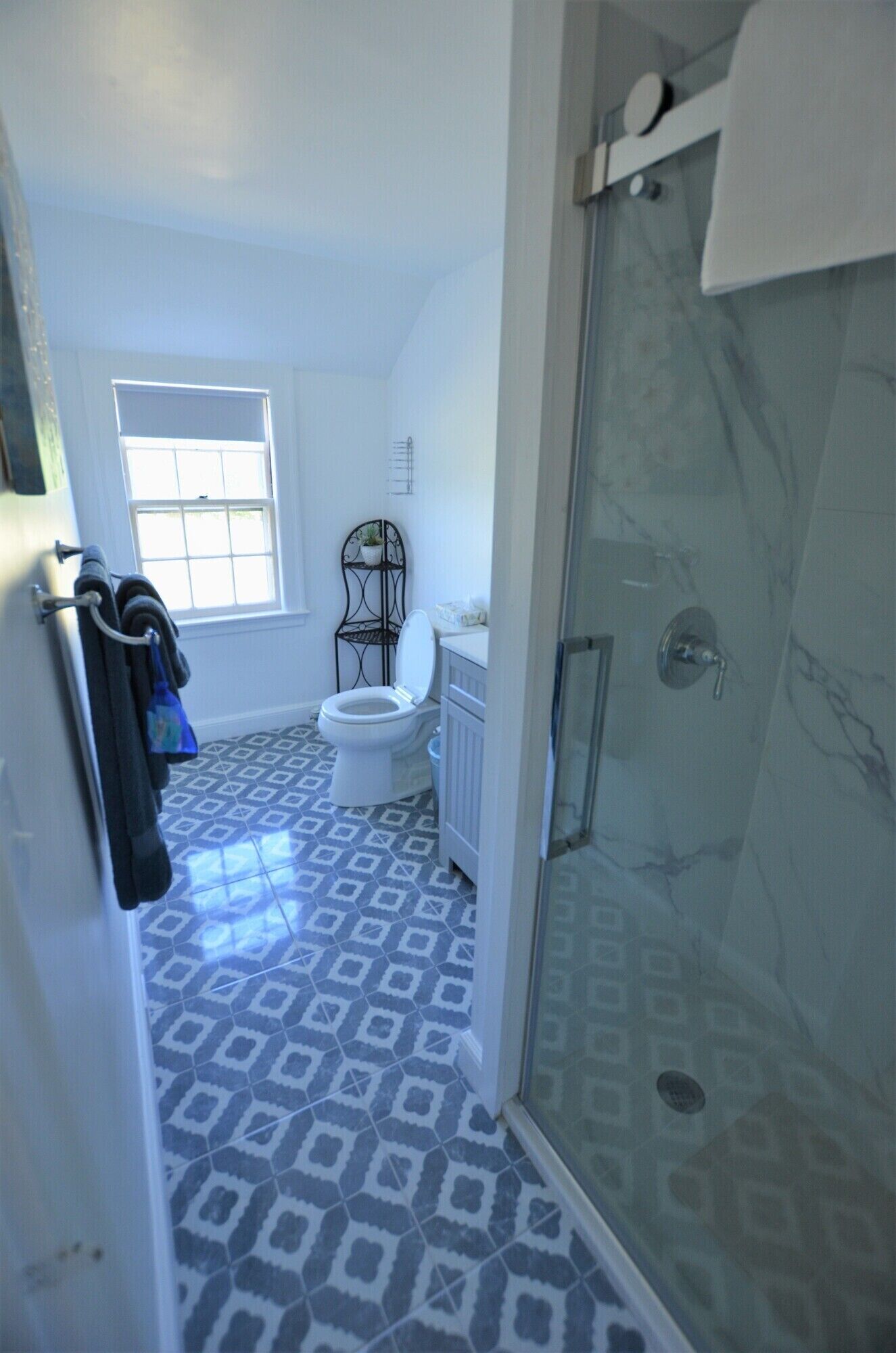 Un baño con piso estampado, inodoro, ducha con puerta de vidrio y ventana que proporciona luz natural.
