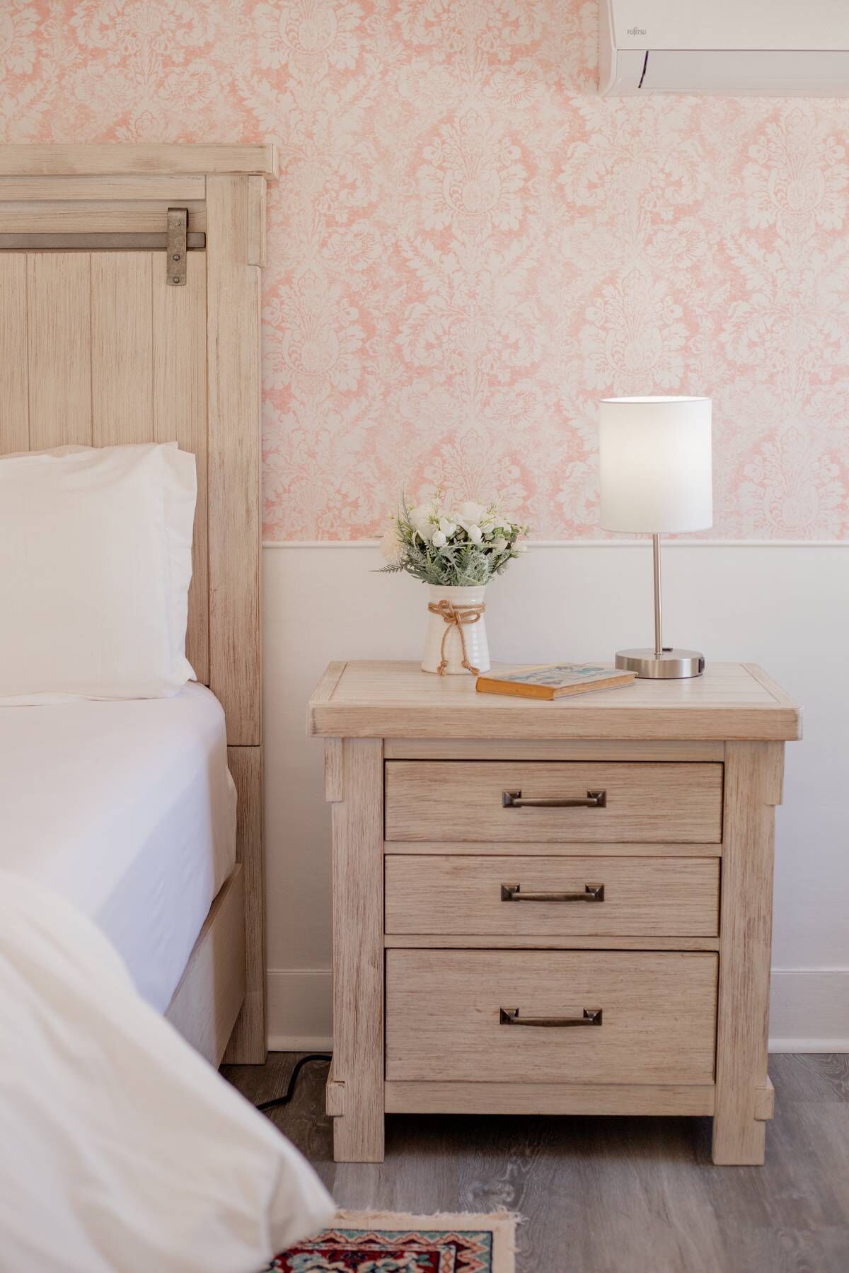 A bedroom with a wooden nightstand, a lamp, and a floral-patterned wallpaper.