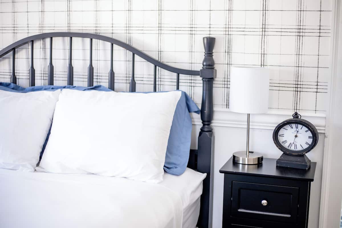 Una cama bien hecha con ropa de cama blanca y azul, un cabecero de metal negro, una mesita de noche con un reloj y una pared con estampado de cuadros.