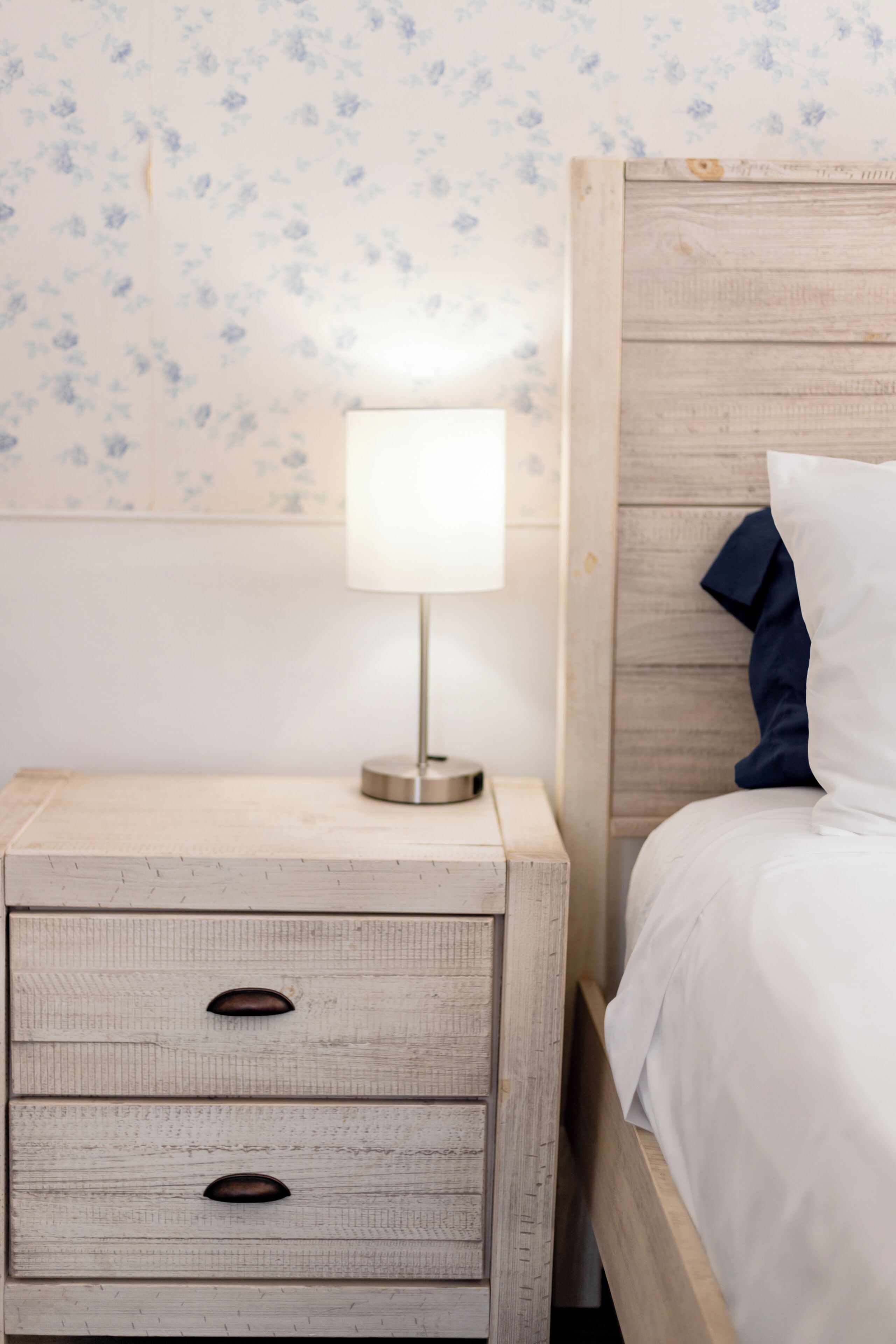 Una mesita de noche de madera con dos cajones y una lámpara, junto a una cama con cabecera de madera y almohada azul.