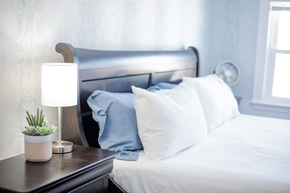 Una cama bien hecha con ropa de cama blanca y azul, una lámpara de noche y una pequeña planta en maceta sobre una mesita de noche de madera oscura.