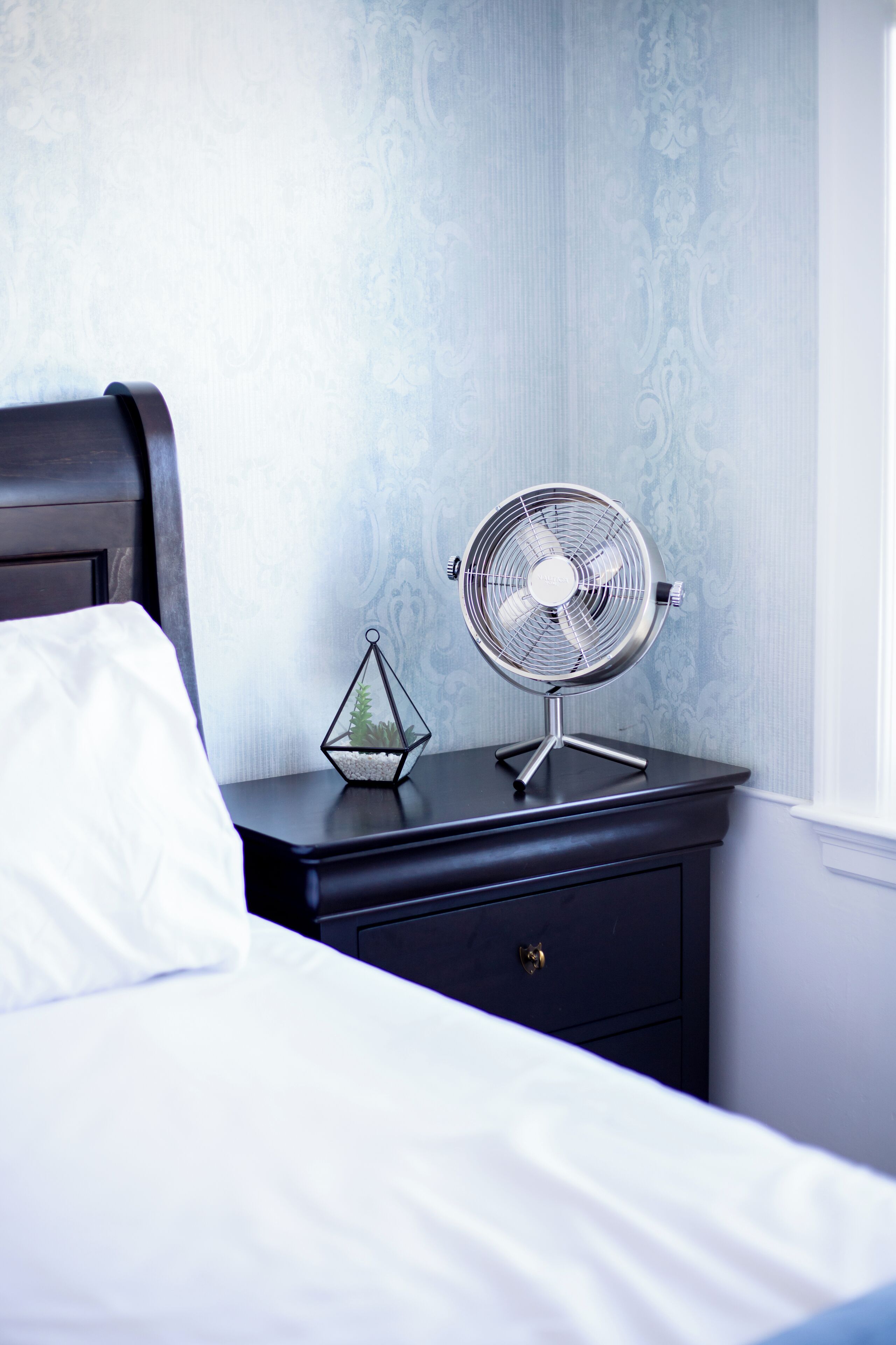 A neatly made bed with white linens, a small bedside table with a fan and a decorative glass container, and a patterned wallpaper background.