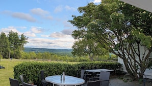 An outdoor patio with a round table and chairs, surrounded by a wooden deck and a view of a green landscape. - Blue Bear Inn (Francestown)