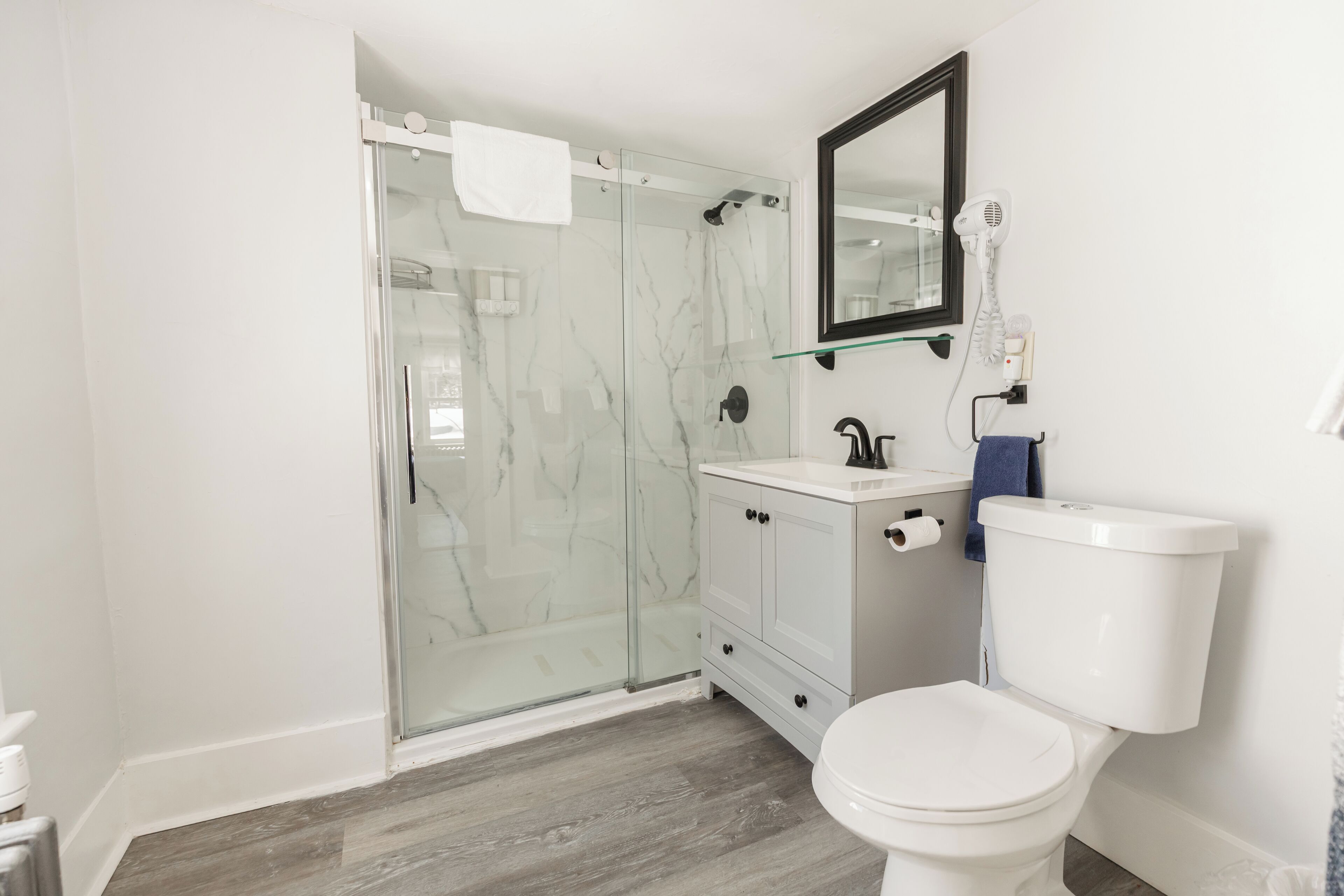 A modern bathroom with a glass shower enclosure, a white toilet, a vanity with a mirror, and a hairdryer mounted on the wall.