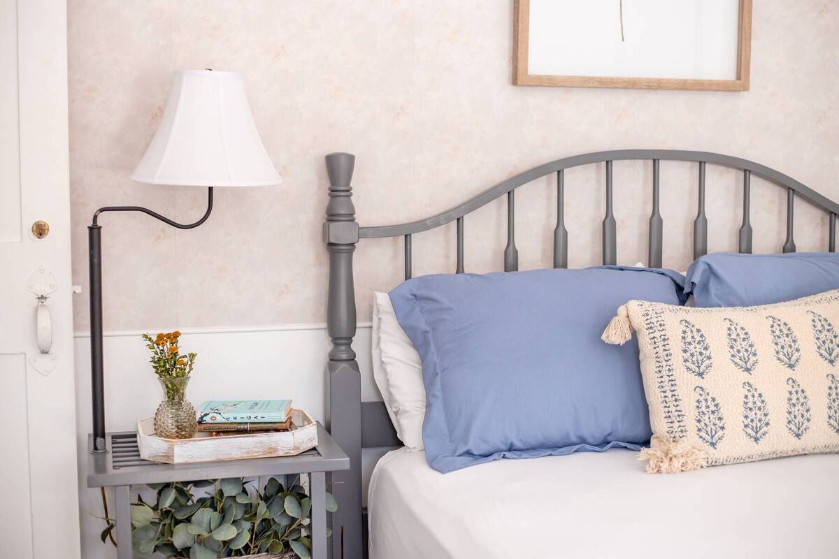 Un dormitorio ordenado con cama, almohadas, mesita de noche con lámpara, un jarrón con flores y un cuadro en la pared.