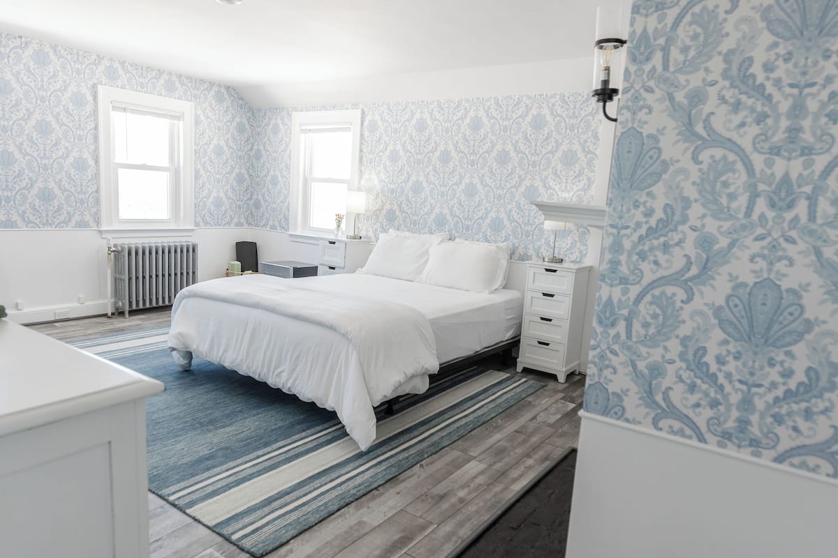 Un dormitorio con cama, mesitas de noche, radiador y papel tapiz estampado.
