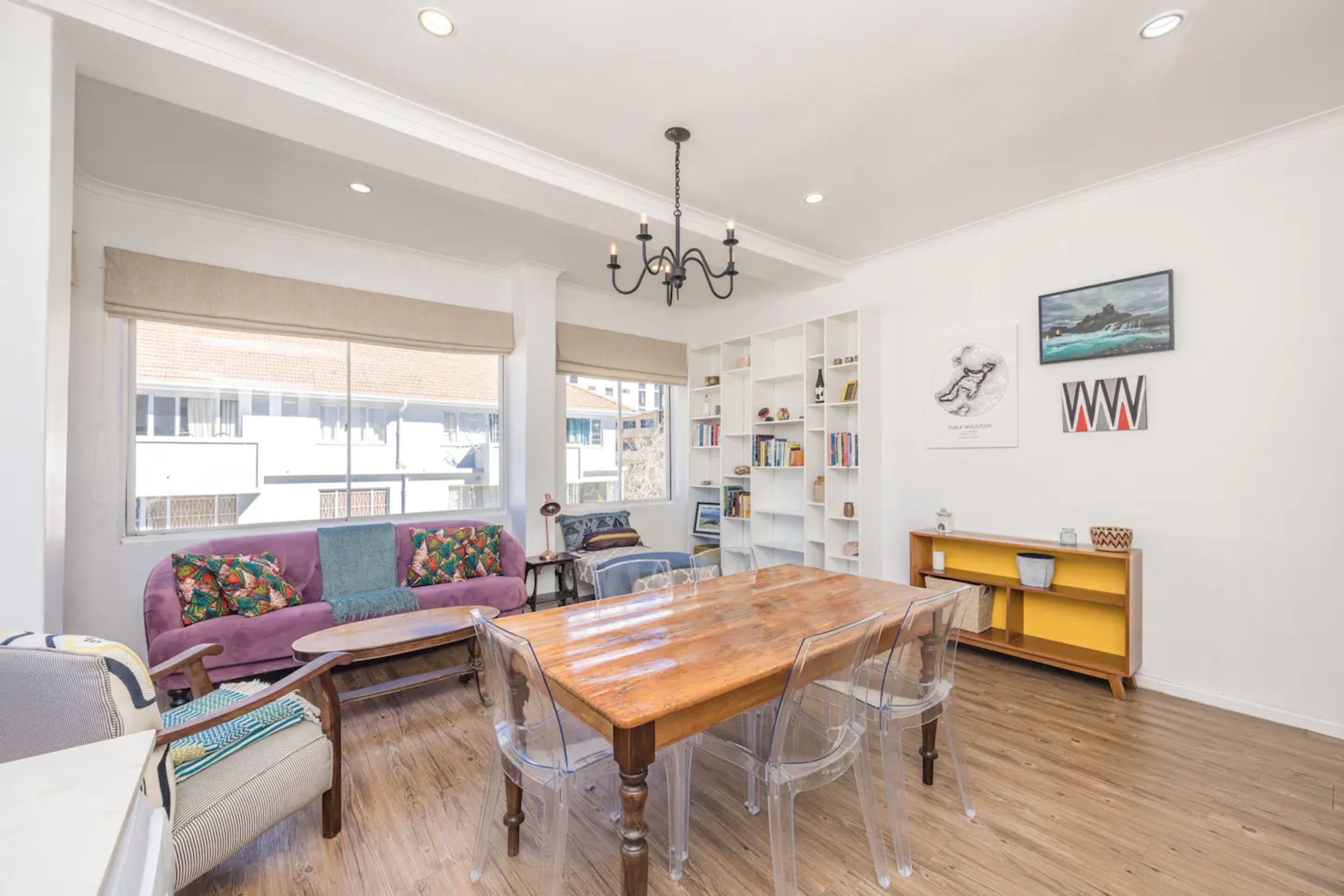 A spacious living area with a wooden dining table, clear plastic chairs, a purple sofa, and a bookshelf.