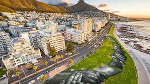 A cityscape with modern buildings, a prominent mountain, and a large mural of hands holding each other on the beach. - Amazing 2 Bedroom Apartment With Mountain Views on Kloof Street (Cape Town)