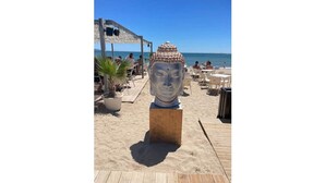 Een strandsetting met een Boeddhabeeld op een houten terras, omringd door ligstoelen en tafels, en een beschut gedeelte met parasols.