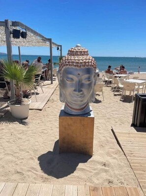Escena de playa con una estatua de Buda sobre una plataforma de madera, rodeada de sillas y mesas, y un área cubierta con sombrillas.