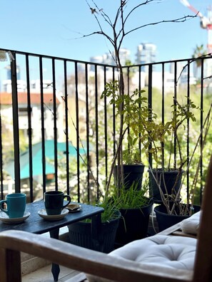 Un balcon donnant sur des immeubles, une table avec deux tasses et des plantes en pot.