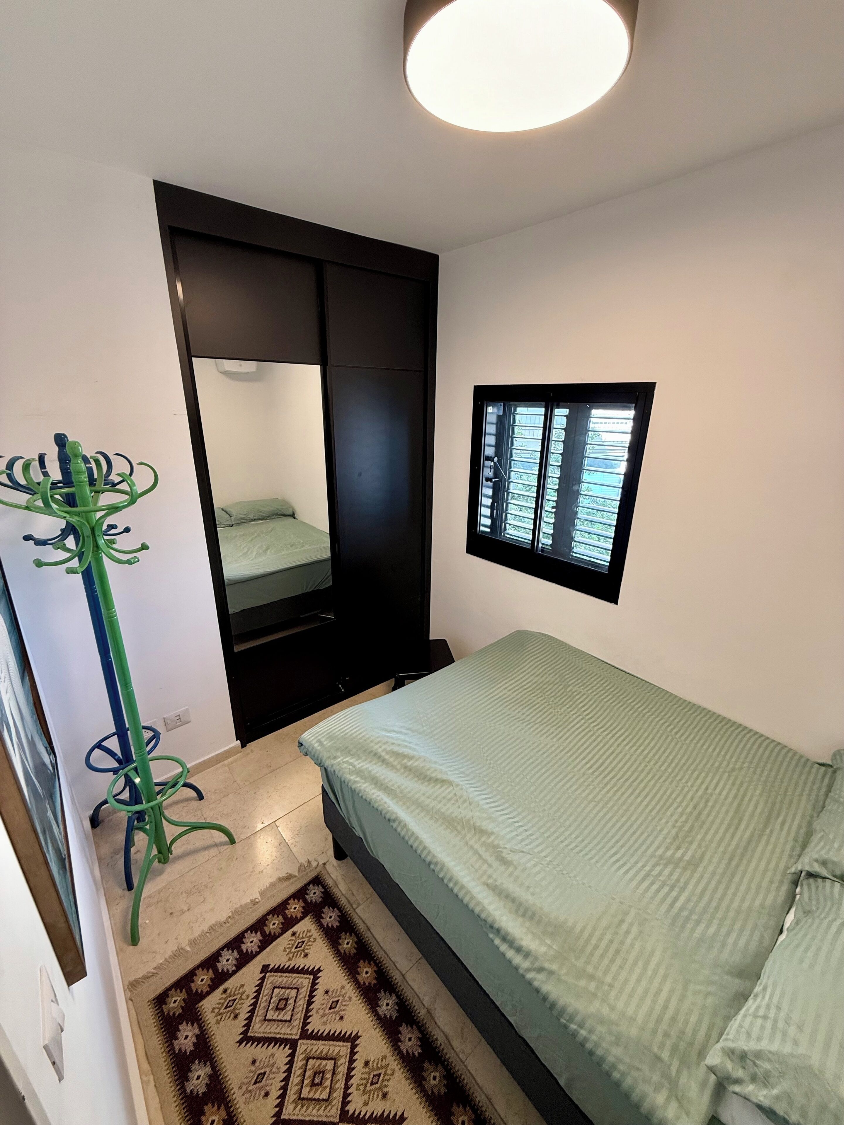 Un dormitorio con una cama, una ventana con persianas, un perchero y una puerta que da a otra habitación.