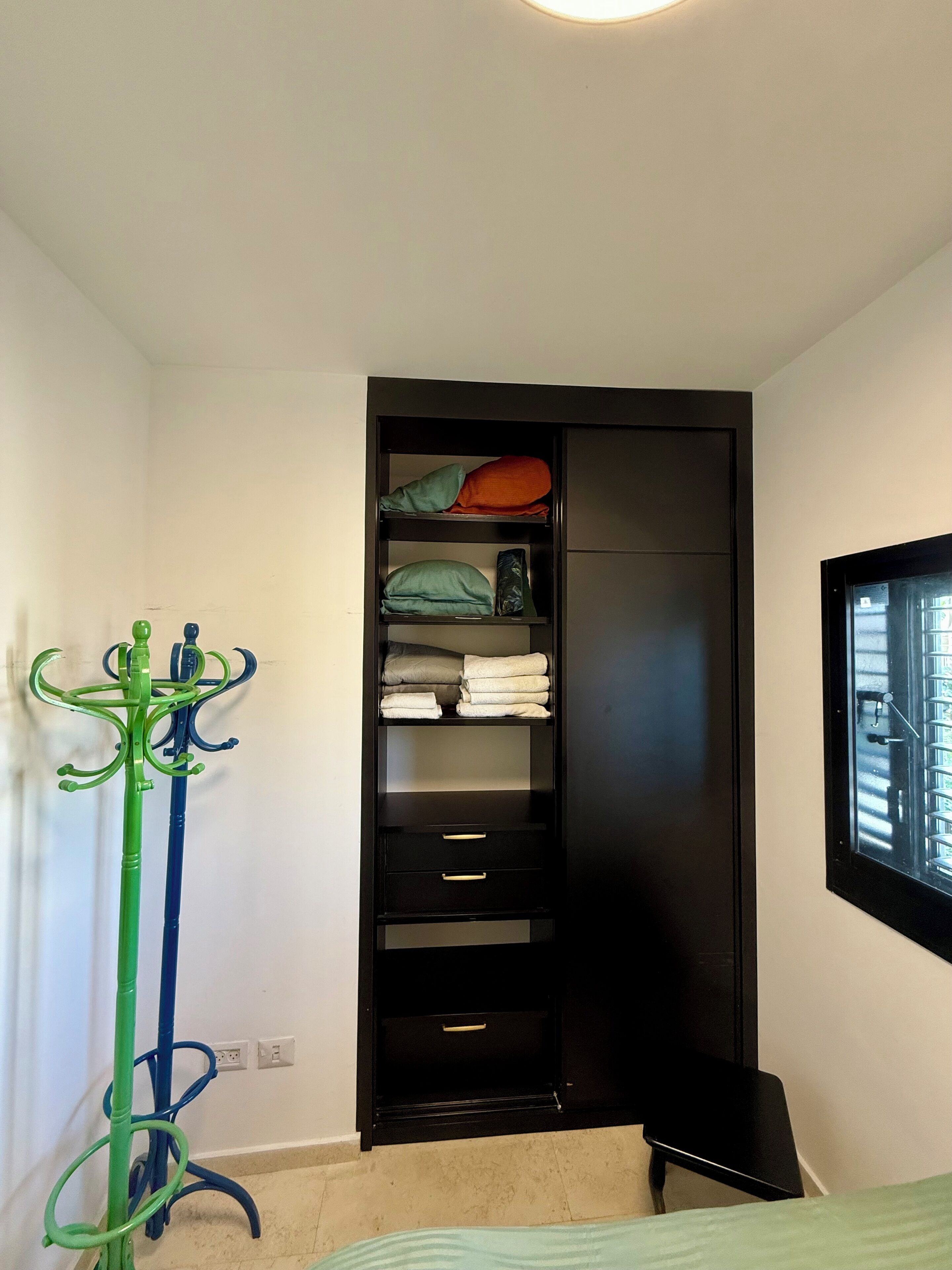 A room with a black wardrobe, a green and blue coat rack, and a bed with a green blanket.