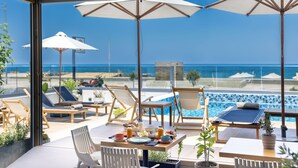 A sunlit outdoor dining area with a pool view, featuring lounge chairs, umbrellas, and a table set for breakfast. - Calla Luxury Seafront Suites (Rethymno)