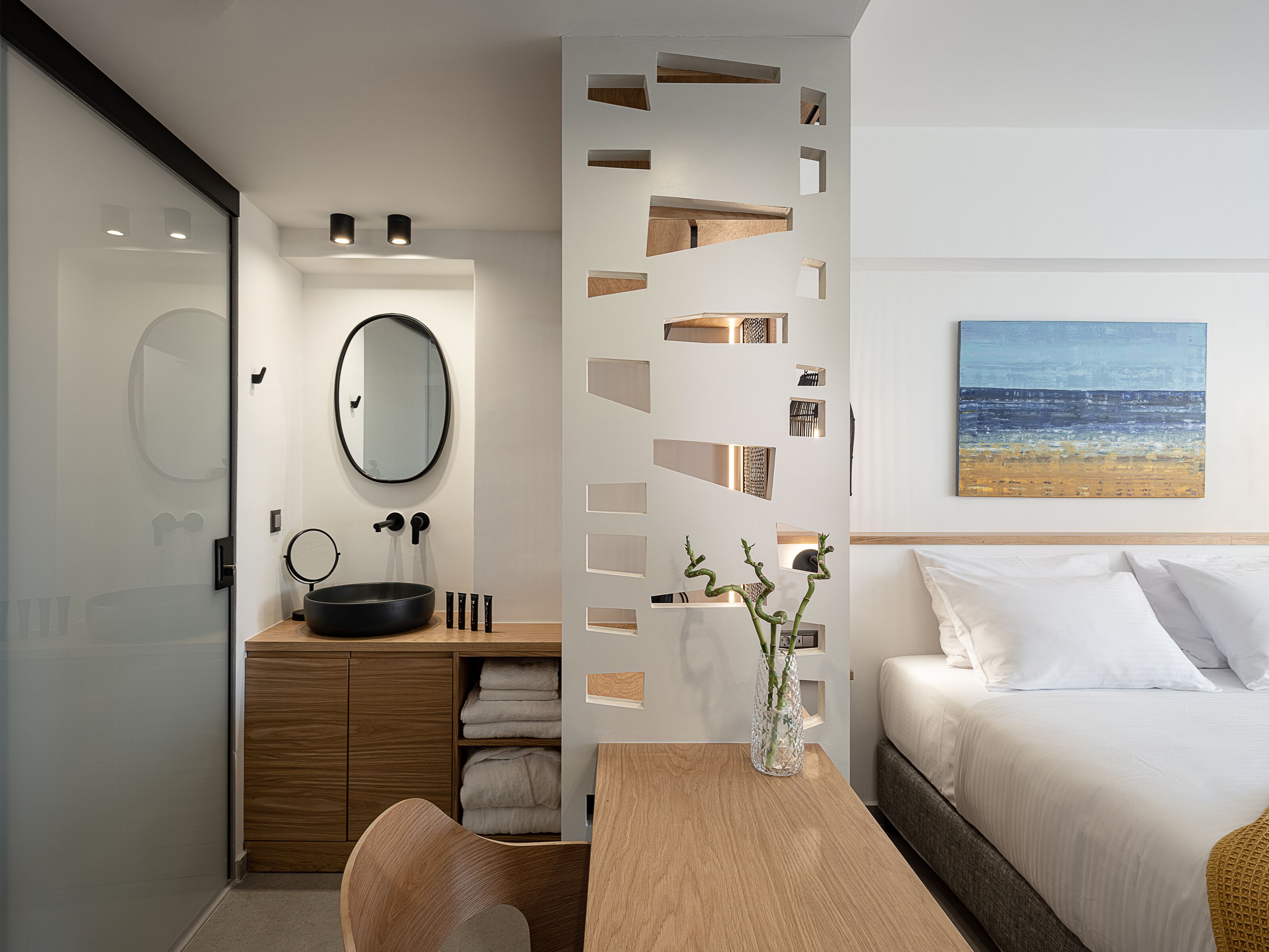 A modern hotel room with a wooden vanity, a round mirror, a wooden table with a vase, a bed with white linens, and a beach painting on the wall.