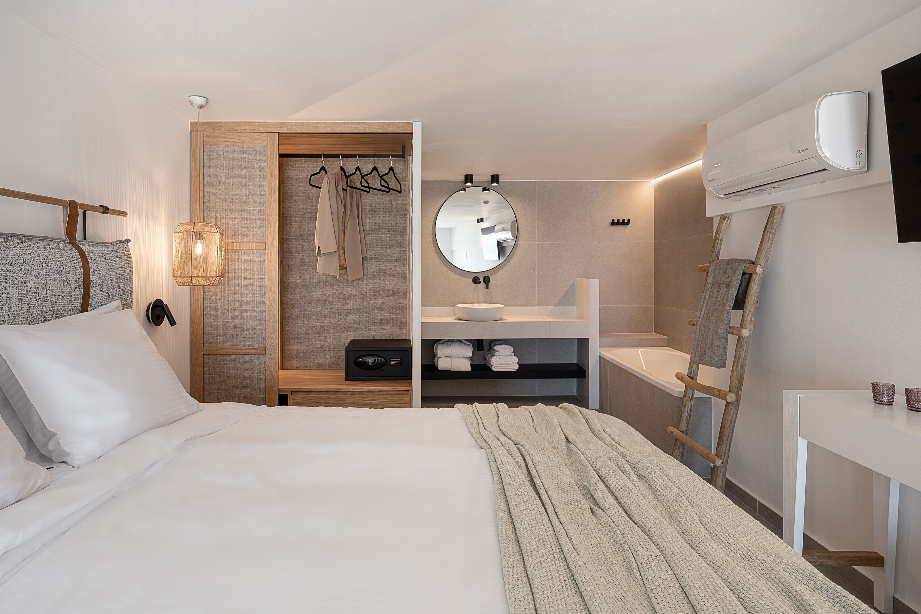 A modern hotel room with a large bed, a bedside table, a mirror, a built-in wardrobe, a vanity with a sink, and an air conditioning unit.