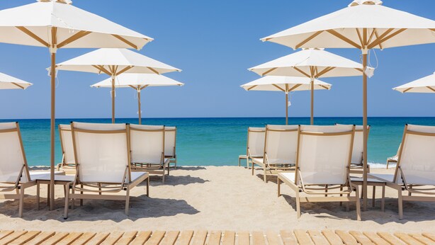 A beach with rows of white sun umbrellas and lounge chairs facing the ocean. - Calla Luxury Seafront Suites (Rethymno)