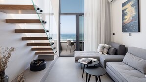 A modern living room with a glass staircase, a sofa, a small table, and a view of the sea. - Calla Luxury Seafront Suites (Rethymno)