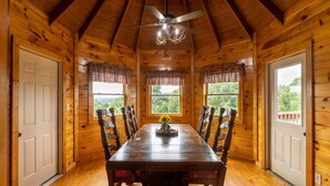 L’intérieur d’un chalet en bois comprend une table à manger, des chaises, un ventilateur de plafond et des fenêtres avec des rideaux.