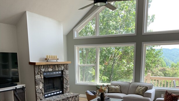 A spacious living room with a stone fireplace, large windows offering a view of trees and mountains, a ceiling fan, and a cozy seating area. - Amazing view from the Family Lake Retreat (Murphy)