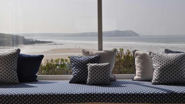 Ein Balkon mit einer gemusterten Couch und Kissen, mit Blick auf einen Strand und eine entfernte Berglandschaft.