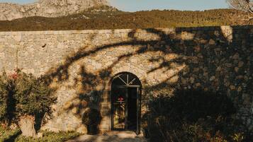 Un edificio de piedra con un arco central, rodeado de follaje y montañas al fondo.