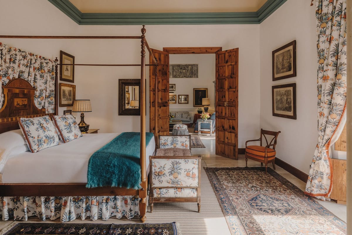 A bedroom with a four-poster bed, a wooden bench, a chair, and a small table. There are multiple framed pictures on the walls and a large mirror.
