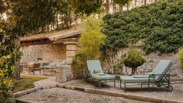 A stone patio with two green lounge chairs and a table, surrounded by greenery and a stone wall.