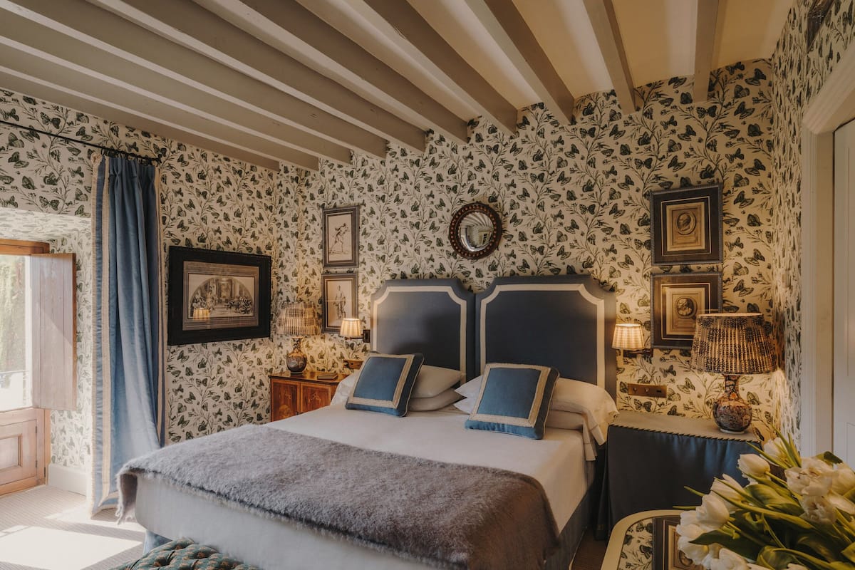 A bedroom with a large bed, a wooden headboard, a nightstand, a lamp, and floral wallpaper.