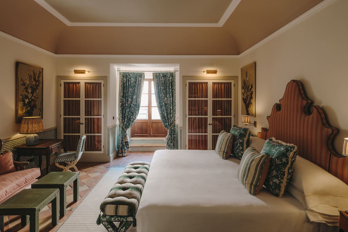 A bedroom with a large bed, a wooden headboard, a bench, a desk, and two chairs. There are two doors leading to a balcony.