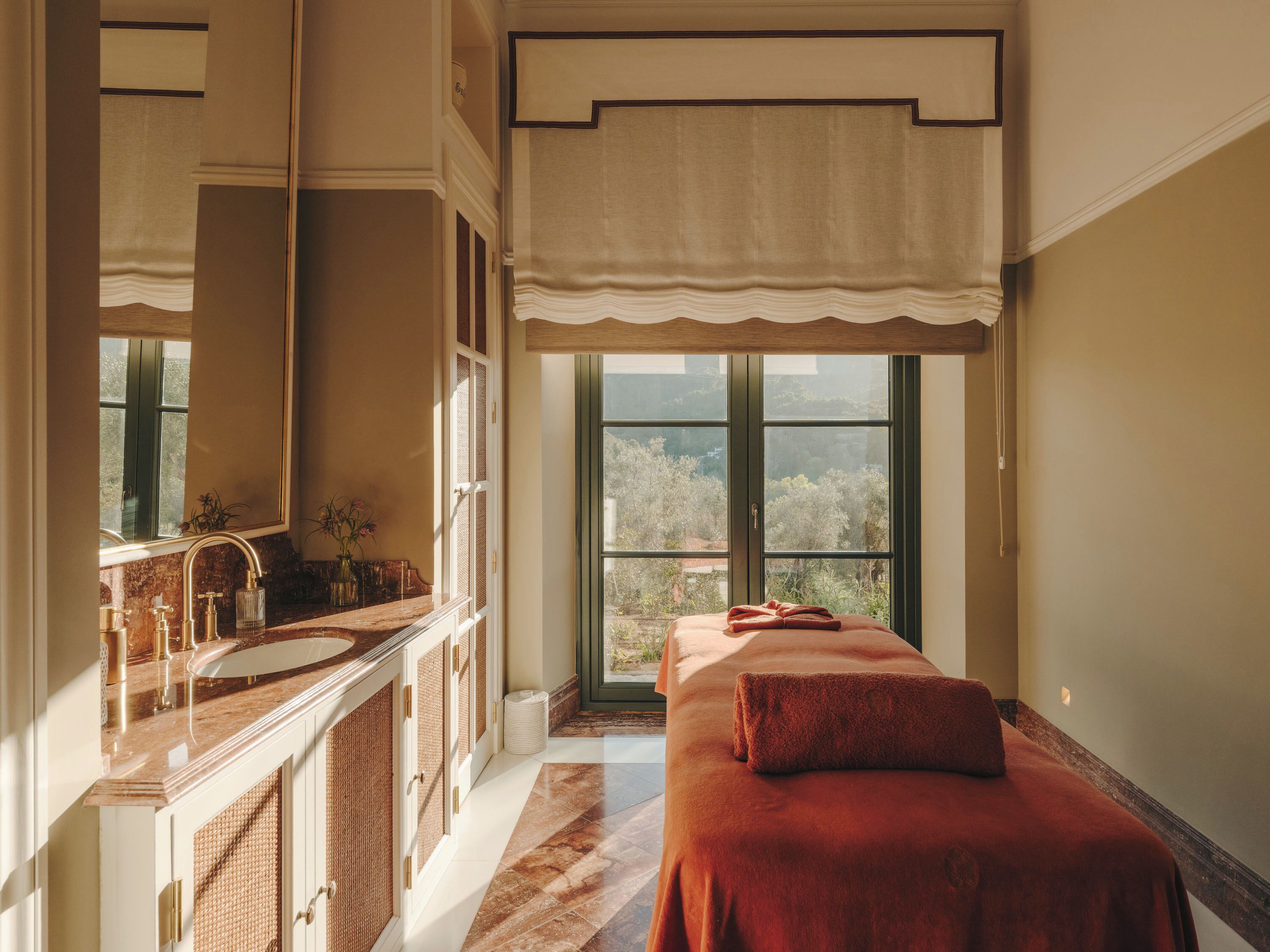 A room with a large window, a bed with a red blanket, a bathroom with a marble sink, and a mirror.