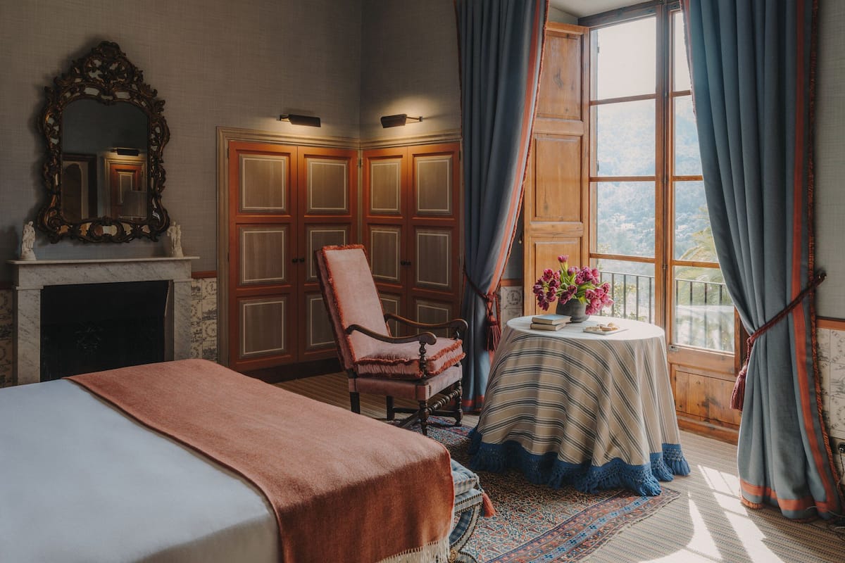A bedroom with a bed, a chair, a table with flowers, a fireplace, and a large window with curtains.