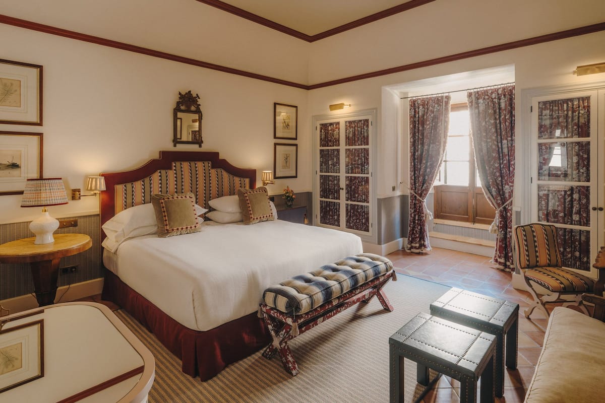 A spacious bedroom with a large bed, a wooden bench, a small table, and a chair. There is a door leading to a balcony with curtains.