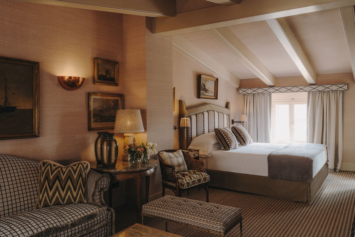 A hotel room with a large bed, a sofa, a small table, and a chair. There are framed pictures on the wall and a lamp on the table.