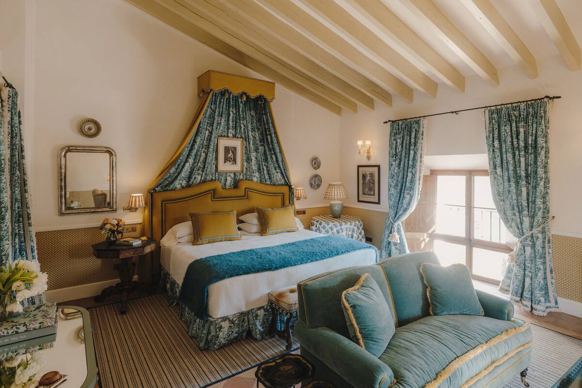 A bedroom with a bed, a sofa, a window with curtains, a mirror, and a bedside table with lamps.