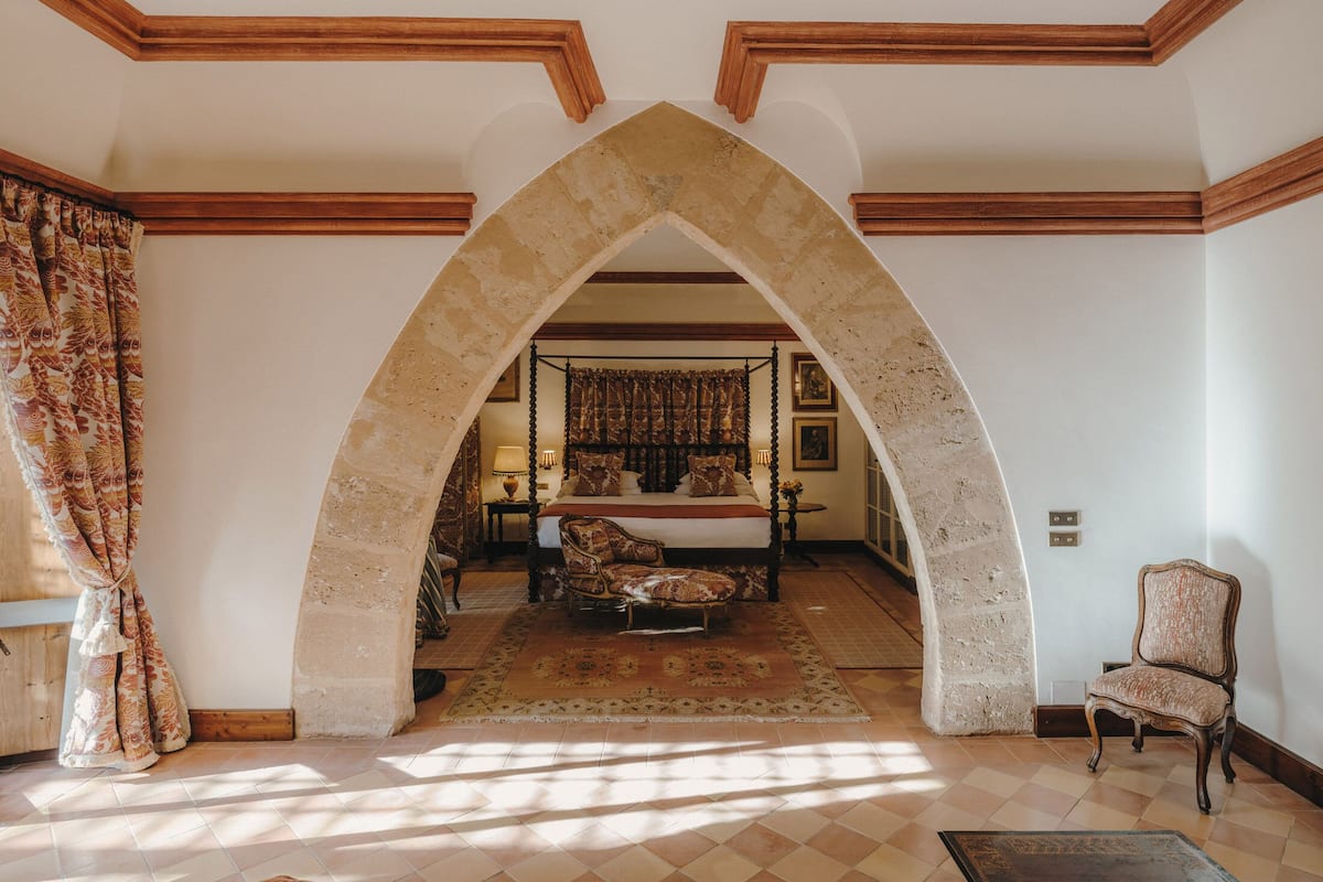 A bedroom with a four-poster bed, a sitting area with a chair, and a view through an arched doorway to another room.