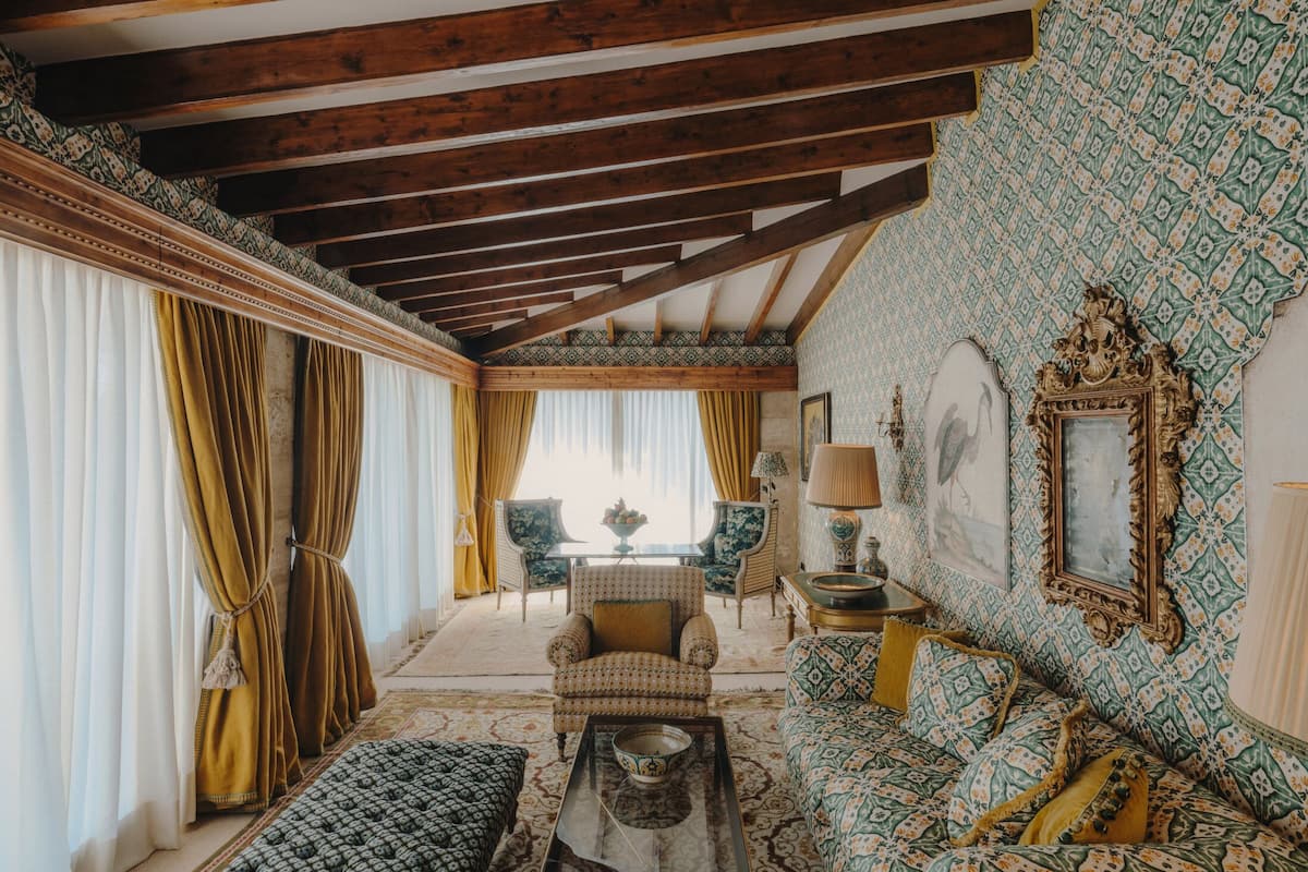 A spacious living room with a high ceiling, wooden beams, patterned wallpaper, and classic furniture.