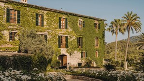 Un edificio con fachada de piedra parcialmente cubierta de hiedra, con varias ventanas con contraventanas, una entrada central y un camino de piedra que conduce a ella.